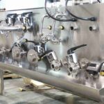 Stainless steel industrial piping system with multiple valves, fittings, and gauges mounted on a panel in a workshop